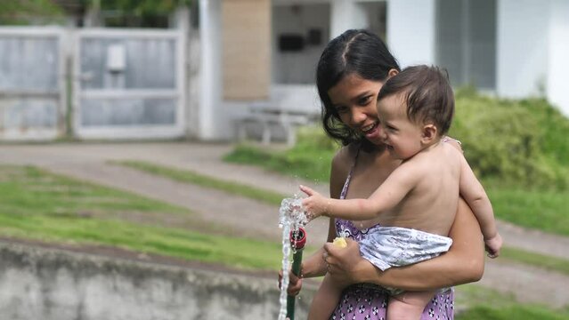 A woman with a child not far from the house holds a hose from which a stream of water pours and shows this process to her small child holding it on his hand. Mom has fun with her 9 month old son. - Powered by Adobe