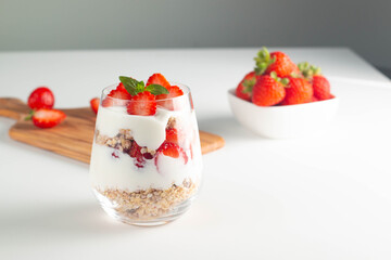 Glass with granola, yogurt and fresh strawberries on white table. Healthy breakfast bowl concept.