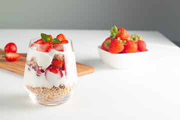 Glass with granola, yogurt and fresh strawberries on white table. Healthy breakfast bowl concept.