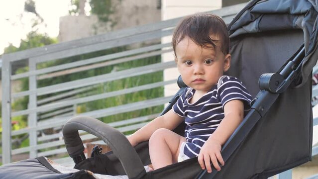 A cute baby 8-10 months old sits in a stroller, thinks about something, stares at it, after which it smiles funny. Carefree childhood of kids who know the world and learn to coexist in it.