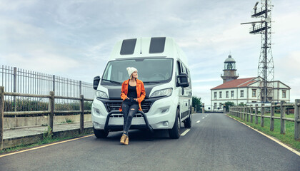 Young woman leaning on camper van on the road with lighthouse in background
