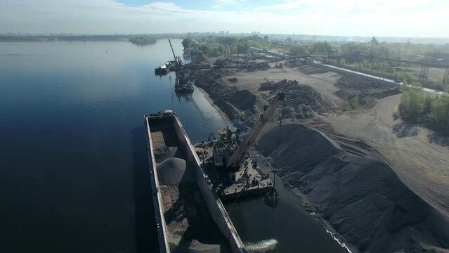 The moment of loading, port crane crushed stone on dry cargo ship. Loading of sand and crushed stone on the river in an industrial area. Orbit aerial panorama.