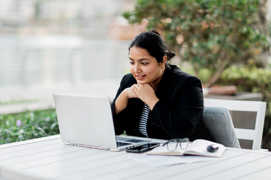 A young and confident Indian Asian woman smiles while sitting and working on her laptop in a stylish cafe on the street on a summer day.