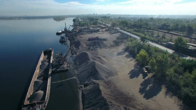 The moment of loading, port crane crushed stone on dry cargo ship. Loading of sand and crushed stone on the river in an industrial area. Orbit aerial panorama.