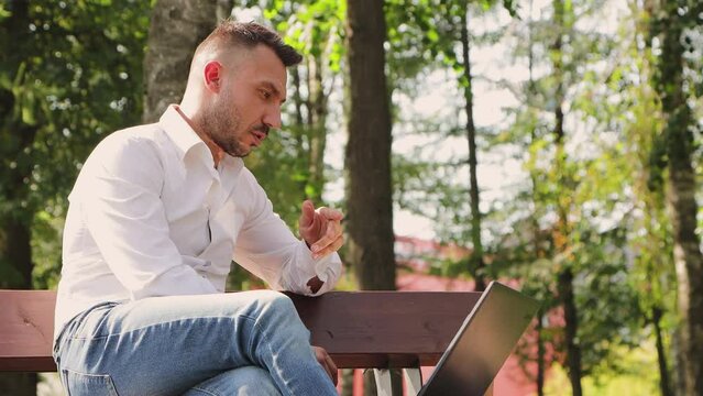 Business man in a white shirt working online with a laptop on a bench. Young man in a city park, hot sunny summer day. Warm soft light, close-up. Against the background of green trees. 4K UHD.