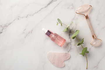 Skincare concept. Top view photo of rose quartz roller gua sha massager glass pink transparent bottle with liquid cosmetics and eucalyptus branch on white marble background with copyspace
