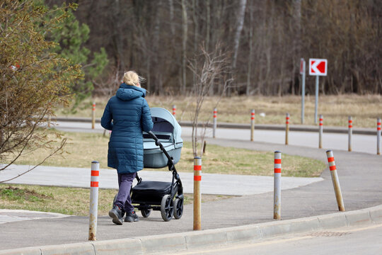 Blonde woman walking with baby pram on a street. Mother in city park at spring