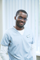 Portrait of smiling black surgeon in scrubs and glasses standing against blinds in office of...