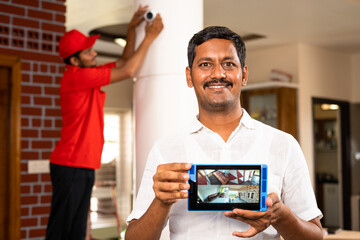 House owner showing cctv camera footage on tablet in front of technician fixing camera at home -...