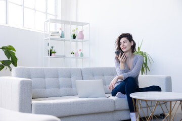 Woman sitting on sofa at home with laptop and have a call on her smartphone while smileing