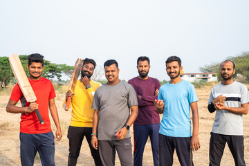 focus on man with yellow t-shirt, Team of cricket players with bat and ball coming and standing by looking at camera on field - concpet of competition, champions and active lifestyle.