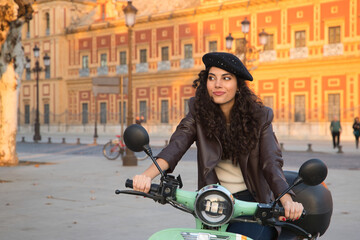 Portrait of a young and beautiful woman, brunette, with curly hair, with leather jacket and black beret, mounted on a rental motorcycle while visiting the city. Concept beauty, fashion, transport.