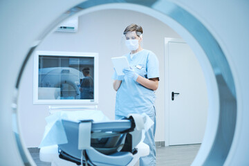 Serious nurse in protective mask and gloves using tablet and reading patients card while preparing MRI scanner for covid patient