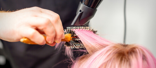 Naklejka premium Drying short pink hair of young caucasian woman with a black hairdryer and black round brush by hands of a male hairdresser in a hair salon, close up