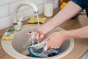 Close up of unrecognizable man in blue shirt washing up white round plate in beige marble kitchen sink under running water from faucet standing in white modern kitchen. Housework, cleaning concept.