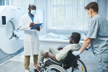 Obraz premium Afro-American doctor in mask with clipboard commenting MRI result to patient in wheelchair pushed by nurse