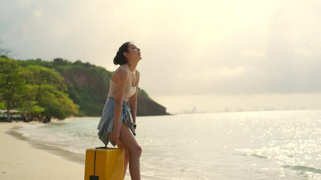 4K Young beautiful Asian woman dragging a suitcase and walking on tropical beach at summer sunset. Cheerful female relax and enjoy outdoor lifestyle on beach holiday vacation and summer travel trip