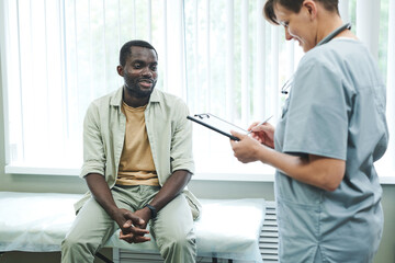 Obraz premium Smiling young nurse in scrubs making notes in clipboard while filling medical records and talking to patient