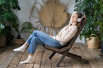 Calm woman sitting in armchair in cozy greenhouse, dreaming, resting, listen to music on wireless...