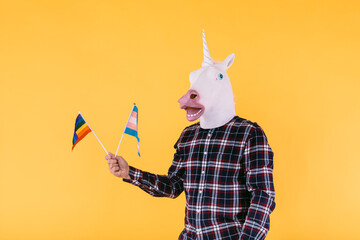 Person dressed in a unicorn mask with a plaid shirt holding a flag of the transsexual and lgtbq community, on a yellow background. Concept of gay pride, transsexuality, gender, binary and lesbian