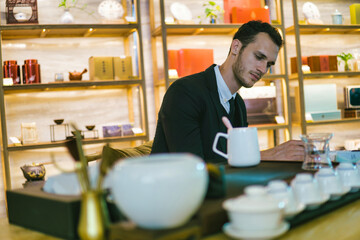 Young handsome caucasian businessman in black suit doing some paperwork. Tea cups and kettle on wooden desk.