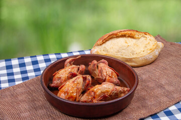 Roasted tiny chicken in clay pan and loaf of white bread lie on rustic table with green outdoor background