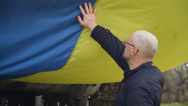 Ukrainian male patriot touches large developing flag of his country on tracked tank during armored invasion. Concept of partnerships and allied countries and nations