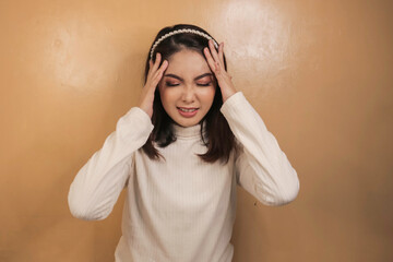 Portrait of young asian woman isolated on orange background suffering from severe headache, pressing fingers to temples, closing eyes to relieve pain with helpless face expression.