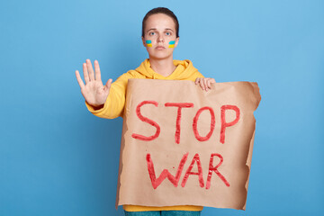 Indoor shot of woman in yellow hoodie with Ukrainian flag on cheeks, posing isolated over blue...
