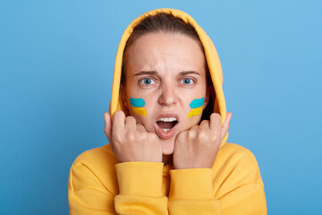 Scared shocked woman in yellow hoodie with Ukrainian flag on cheeks, posing isolated over blue background, looking at camera with frowning face, expressing negative emotions, does not believe. - Powered by Adobe