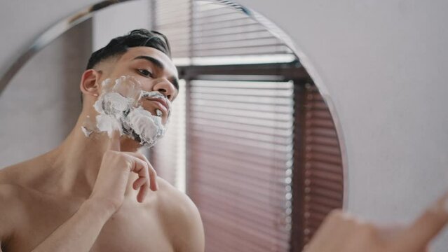 Portrait male reflection in mirror naked man brunette brutal guy in shower smears face with white foam soap shaving gel masculine cosmetics getting ready for procedure to shave daily facial skin care