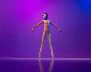 Fototapeta na wymiar Studio shot of beautiful school age girl, ballet dancer dancing isolated on purple background in neon light. Art, grace, beauty, ballet school concept