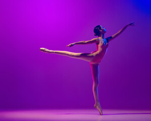 One flexible young female ballet dancer, teen in stage outfit and pointes dancing isolated on purple background in neon light. Art, grace, beauty, ballet school concept