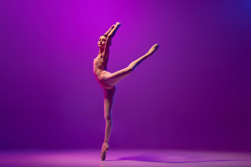 Studio shot of beautiful school age girl, ballet dancer dancing isolated on purple background in...