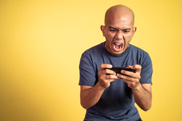 Bald asian man shouting with mouth open while playing game using smartphone on isolated background