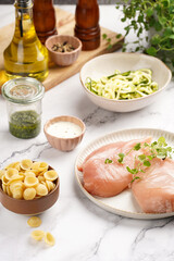 Fresh ingredients for preparing a warm salad on marble surface: chicken breasts, heavy cream, zucchini noodles, italian pasta orecchiette, herbs and sauces