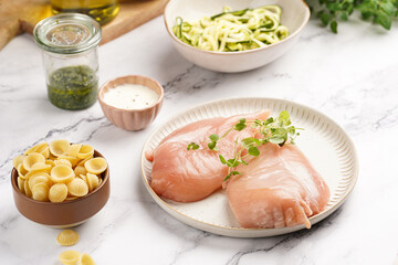 Fresh ingredients for preparing a warm salad on marble surface: chicken breasts, heavy cream, zucchini noodles, italian pasta orecchiette, herbs and sauces