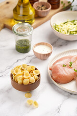Fresh ingredients for preparing a warm salad on marble surface: chicken breasts, heavy cream, zucchini noodles, italian pasta orecchiette, herbs and sauces