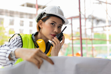 Asian engineer or Young woman Architect put on a helmet for safety and look at Blueprint for Inspect Building factory Construction Site and use walkie talkie for a talk with a contractor.