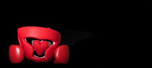 On Valentine's Day, red boxing gloves hug a protective helmet in which lies a heart made of red fabric as a symbol of the holiday. Studio photo on black banner background.