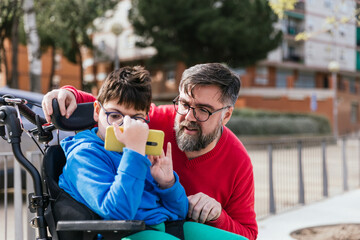 Disabled child in wheelchair watching something on the mobile phone with his father.