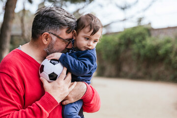 Man holds his little son and a soccer ball while playing together outdoors in the park.