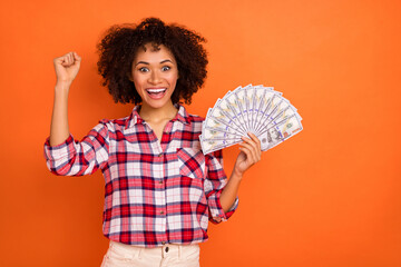 Photo of lucky funny young woman wear plaid shirt rising fist money fan empty space isolated orange color background