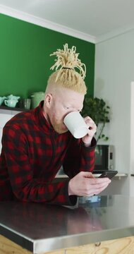Vertical shot of an Albino man leaning over a kitchen isle, drinking coffee and using his smartphone