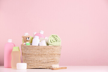 Baby cosmetic products, bath accessories and toy in wicker basket on white table against pink...