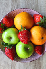 Pink bowl filled with fresh apples, oranges and strawberries on wooden table. Flat lay.