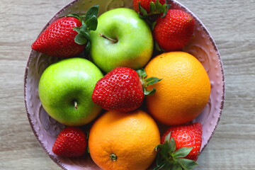 Pink bowl filled with fresh apples, oranges and strawberries on wooden table. Flat lay.