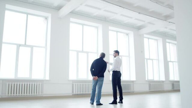 Realtor and client are standing and discussing lease agreement front of windows at office space, back view. Realtor with businessman are standing front of windows in an office space