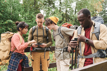 Group of young hikers with backacks using gps on phone while discussing route on forest bridge