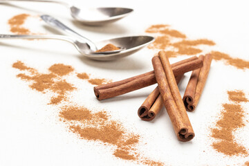 Cinnamon sticks among ground cinnamon, metal spoons on table.